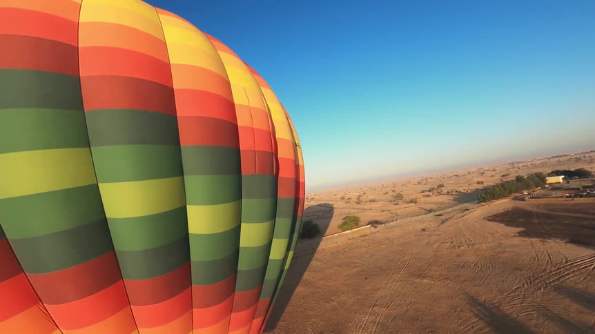 A vibrant, multi-colored hot air balloon ascends over a vast desert landscape under a clear blue sky
