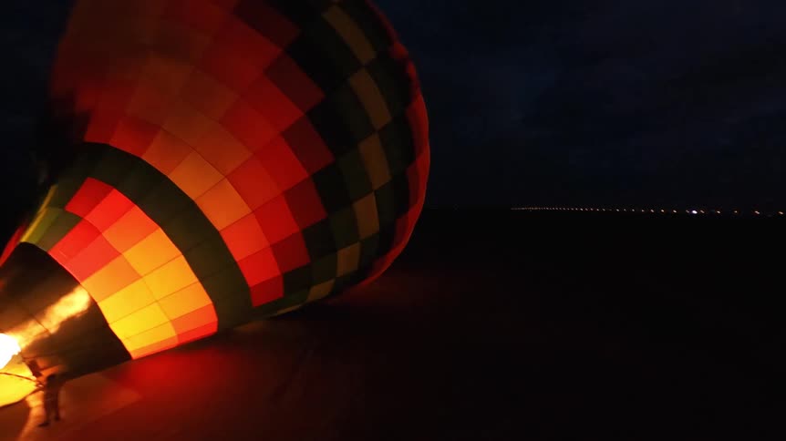 Aerial footage captures a hot air balloon being inflated with a large flame in the dark desert before a sunrise launch