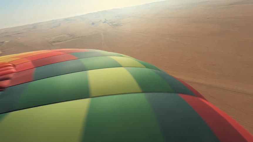 A point of view from a colorful hot air balloon as it ascends and floats over a vast desert landscape with sand dunes