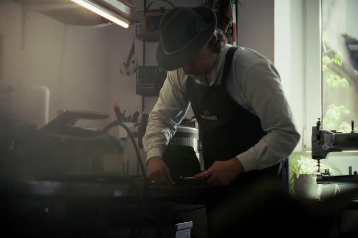 An artisan hat maker in a traditional outfit meticulously brushes and shapes a felt hat in his workshop