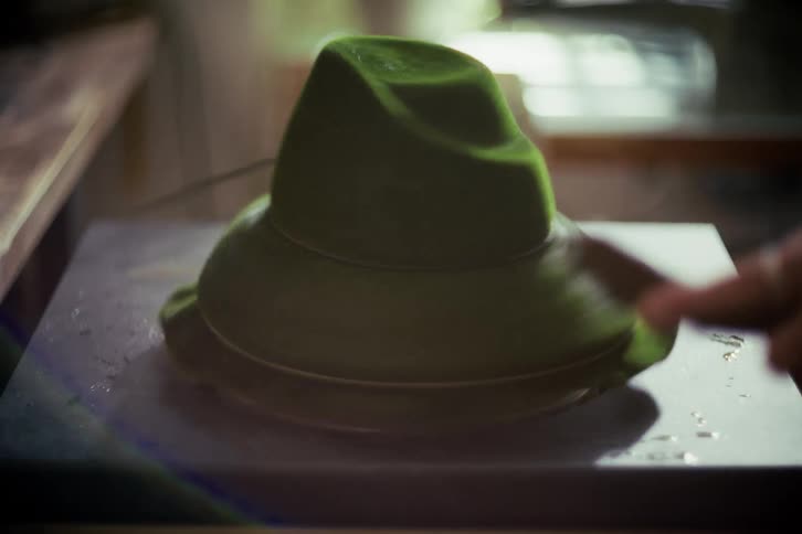 A skilled hatmaker uses a traditional wooden tool to meticulously shape a spinning green felt hat in a workshop