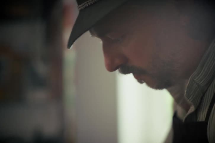 Side profile close-up of a craftsman in a hat concentrating on his work, with a bright light flaring in the background