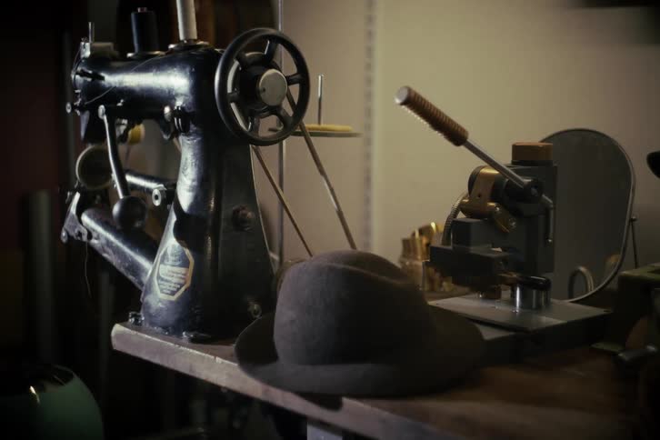 A vintage sewing machine and a classic felt hat rest on a workbench in a dimly lit workshop