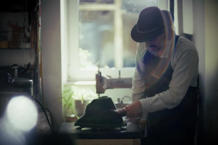 An artisan hatter meticulously brushes a traditional felt hat on a spinning block in his sunlit workshop