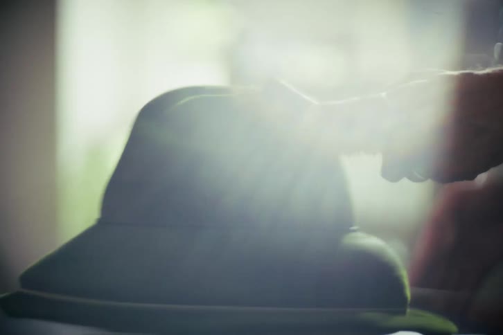 An artisan meticulously brushes a spinning green felt fedora hat in a sunlit workshop