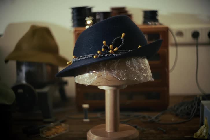 A classic dark blue felt fedora with a decorative cord band sits on a wooden stand in an artisan's workshop