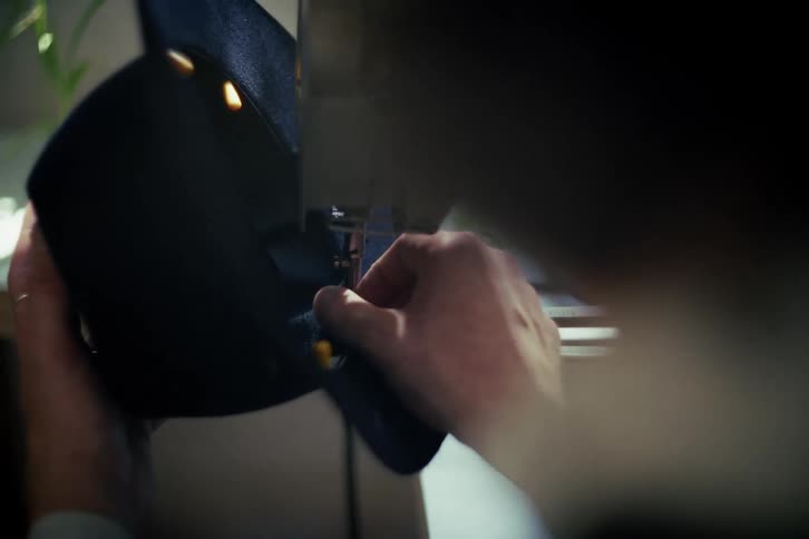 A close-up, over-the-shoulder view of a tailor carefully stitching dark blue fabric on a sewing machine