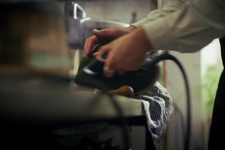 An artisan uses a steam iron to meticulously shape the brim of a green felt hat in a dimly lit workshop