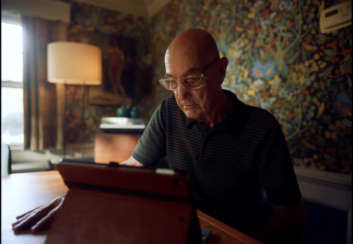 A bald senior man wearing glasses intently reads from a tablet computer while seated at a table in his home