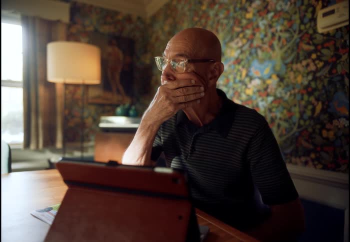 A contemplative senior man sits alone at a table, reacting with concern and sadness to what he sees on his tablet
