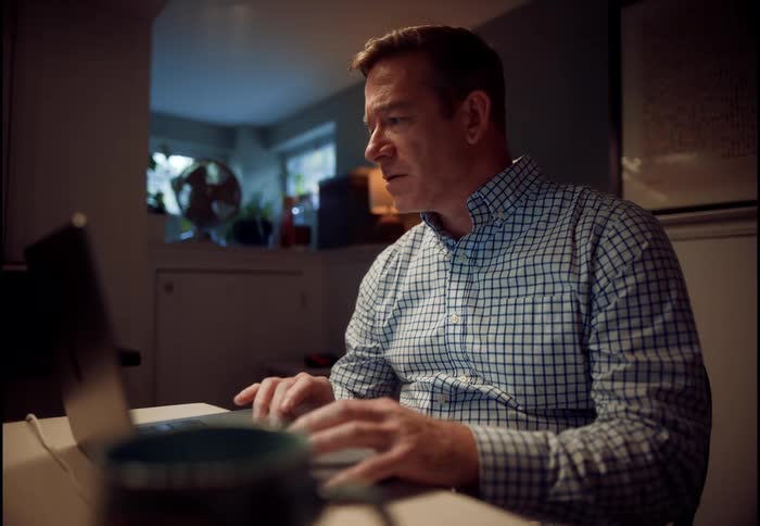 A serious middle-aged man in a plaid shirt works from home late at night, engaging in a video call on his laptop