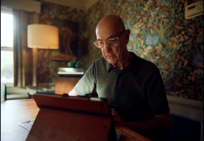 An elderly man with glasses sits at his dining room table using a tablet computer, then looks thoughtfully out a window
