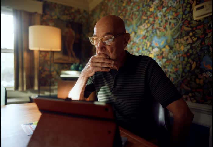 An emotional senior man sits at a dining table and reacts with concern while looking at his tablet computer