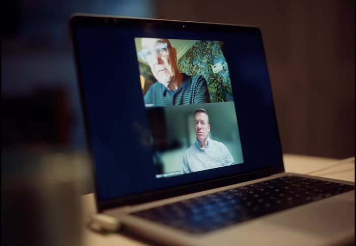 A laptop screen displays a video call between an adult son and his elderly father as they talk in a dimly lit room