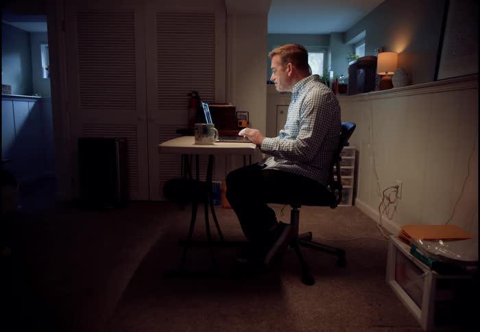 A man on a video call from his dark home office, illuminated by the glow of his laptop screen