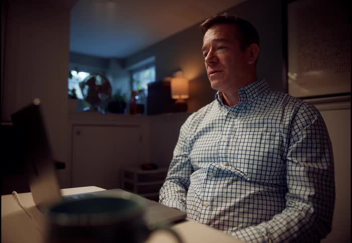 A middle-aged man in a plaid shirt engages in an expressive video conference call from his home office at night