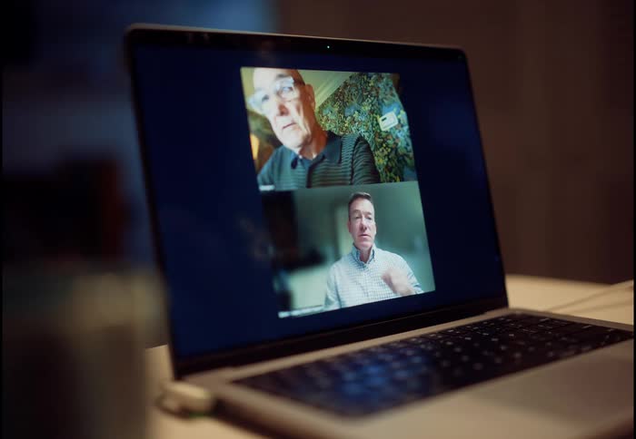 A close-up on a laptop screen displays a video conference call between an older man and a middle-aged man