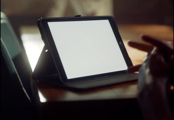 A digital tablet with a blank white screen sits on a wooden desk as a person's hands gesture and tap nearby
