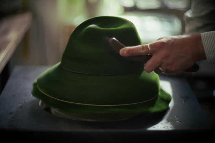 A milliner carefully brushes a green felt fedora spinning on a turntable in a workshop