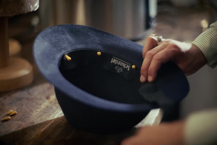 A skilled hat maker carefully uses pins to secure the interior sweatband of a dark blue felt hat in a workshop