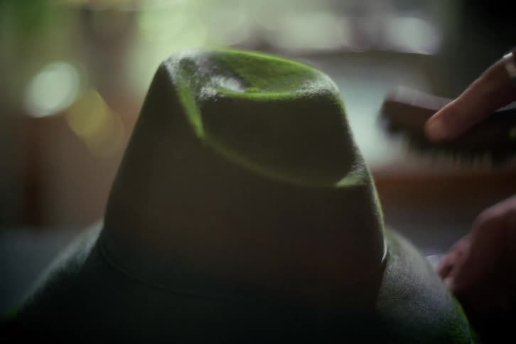 A person carefully brushes a dark green felt fedora hat with a small brush in a softly lit, out-of-focus setting