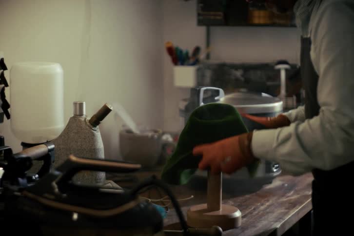A craftsman uses steam to soften a green felt hat before carefully shaping it on a wooden block in his workshop