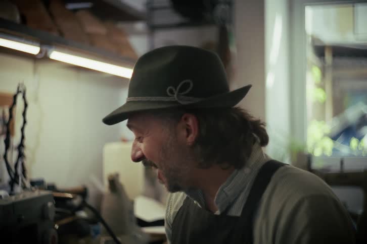 A cheerful artisan hatter wearing a traditional hat smiles and laughs while working in his studio