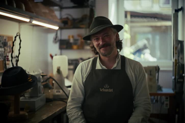A friendly, smiling hatter wearing an apron and hat poses for a portrait in his well-lit workshop