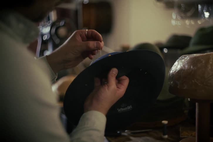 Close-up of a milliner's hands as he carefully hand-sews the interior band of a classic felt hat in his workshop