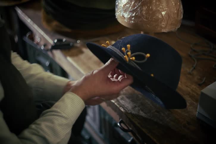 A skilled artisan meticulously hand-sews a decorative band onto a dark felt hat in a traditional workshop