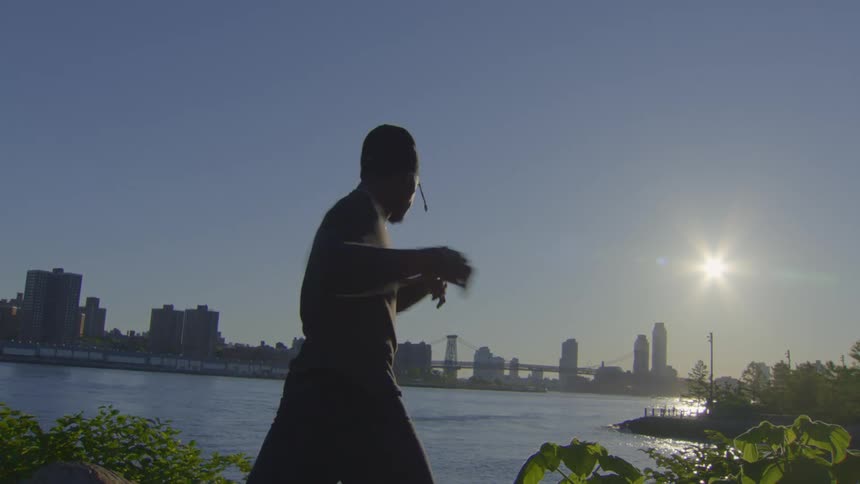 A man in silhouette practices a slow-motion martial art by the river as the sun rises over the city skyline