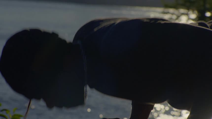 Close-up silhouette of a man doing push-ups with the setting sun reflecting on the water behind him