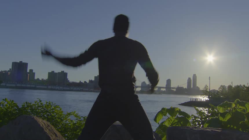 A silhouetted man performs side stretches by a river at sunset with a city skyline and bridge in the background