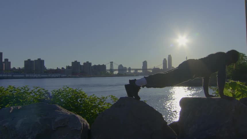 A determined man is silhouetted by the sun as he performs push-ups on rocks with a city skyline across the water