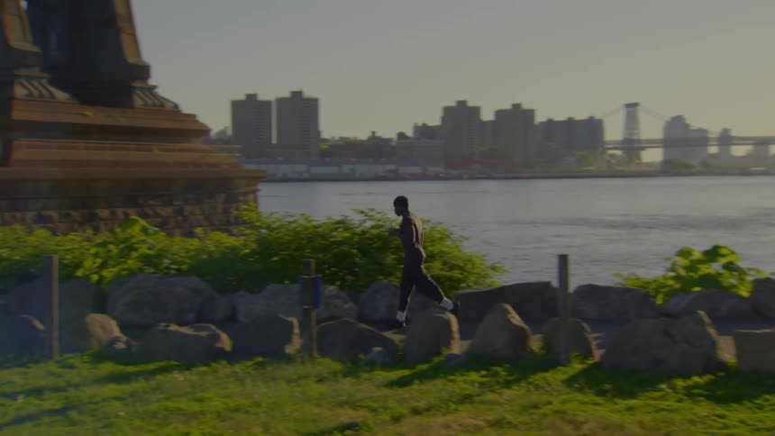 A man jogs in slow motion along a riverside park with the New York City skyline and bridge in the background
