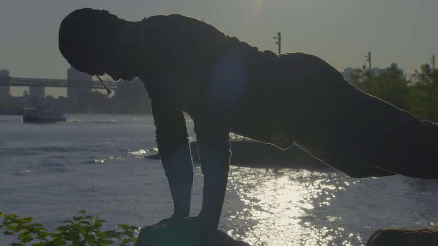A silhouetted man holds a challenging yoga pose in a city park at sunset, with the skyline and river in the background