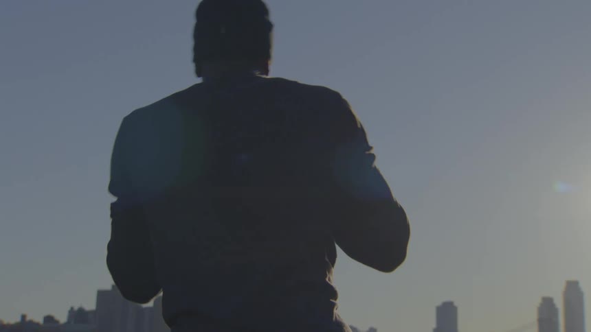 A silhouetted man in slow motion prepares for his morning workout while looking out at the city skyline at sunrise
