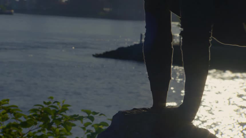 A person's silhouette is captured as they perform an impressive yoga arm balance on a rock overlooking a sunlit river