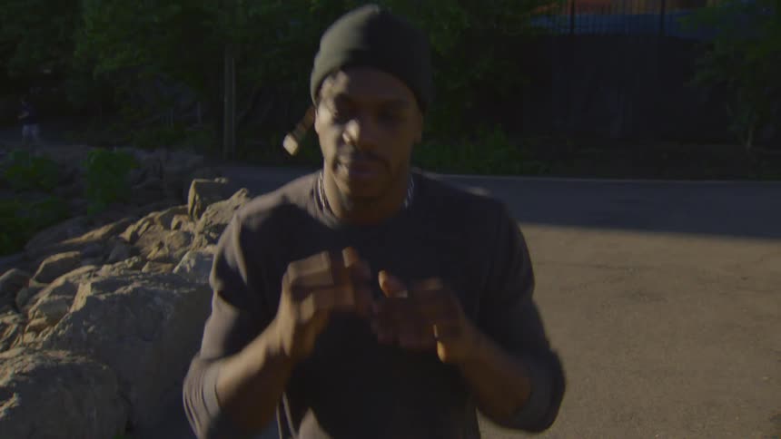 A determined man in a beanie jogs in slow motion on a paved park path during golden hour