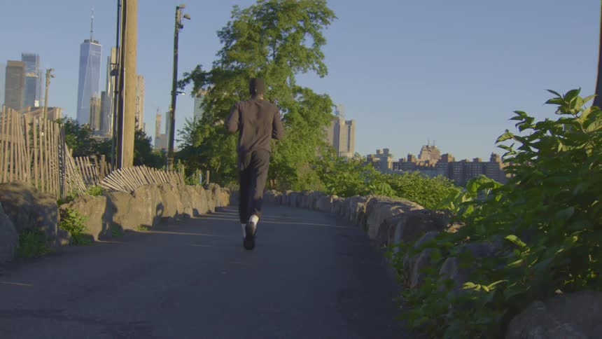 A man jogs on a paved path in a city park with the New York City skyline in the background at sunrise