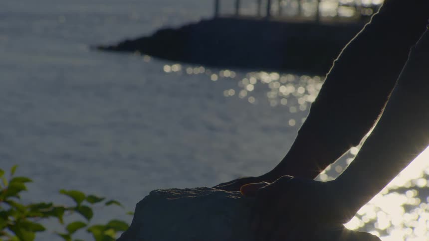 A person's hands rest gently on a rock, silhouetted against a body of water with sunlight shimmering on the surface