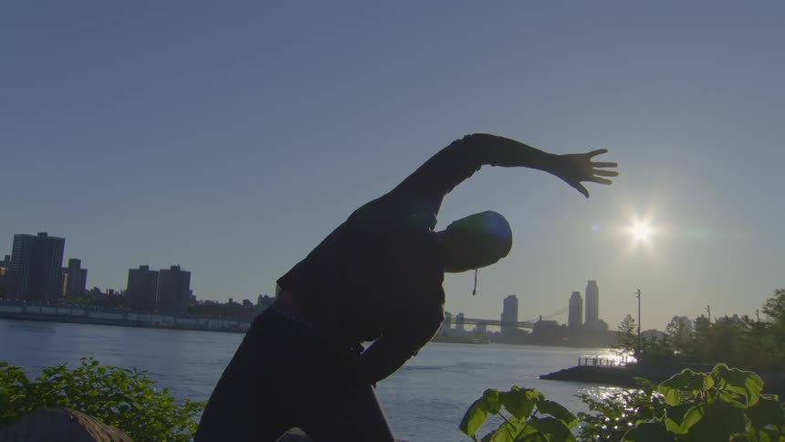 A silhouetted man performs side stretches in a park overlooking a river and city skyline at sunrise