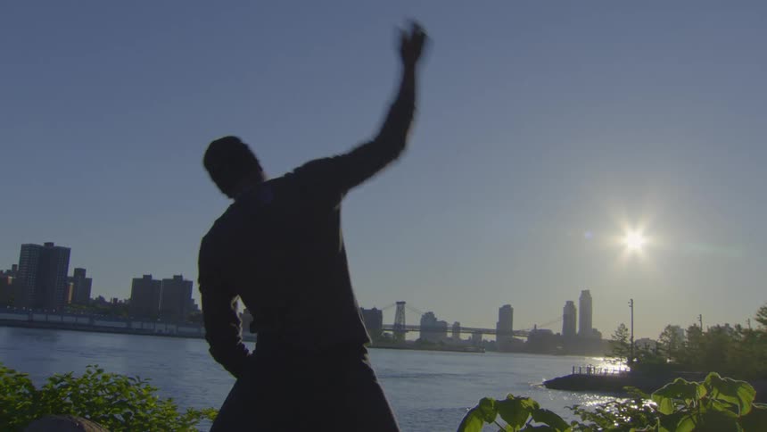 A silhouetted man performs slow-motion stretching exercises by the river at sunrise with the city skyline in the background