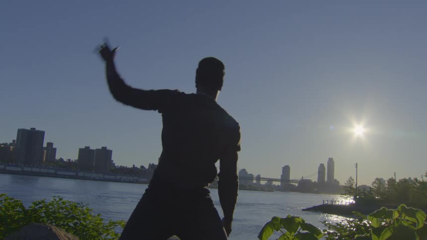 A man in silhouette practices Tai Chi and stretching by the river at sunrise, with the city skyline in the background