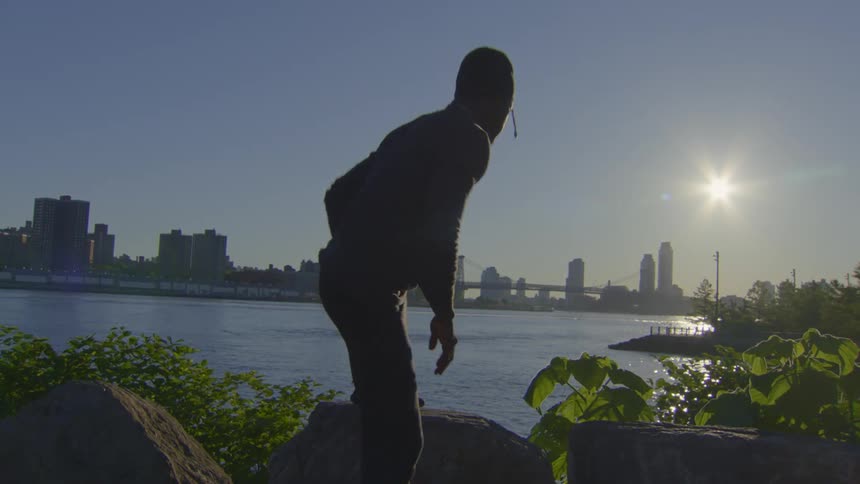 Silhouette of a man stretching on rocks by the river with the city skyline in the background during sunset