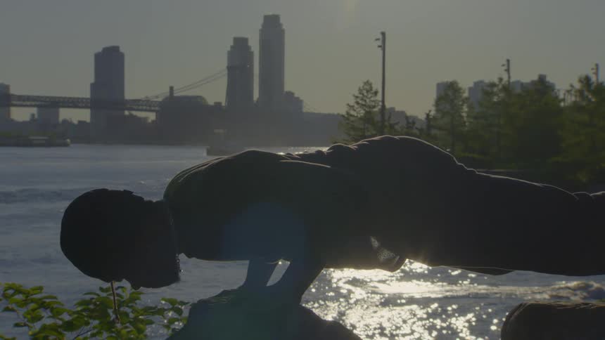 A man performs a slow-motion push-up, silhouetted against the sun rising over a city river