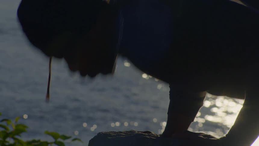 Close up of a silhouetted person doing pushups on a rock overlooking a body of water at sunset