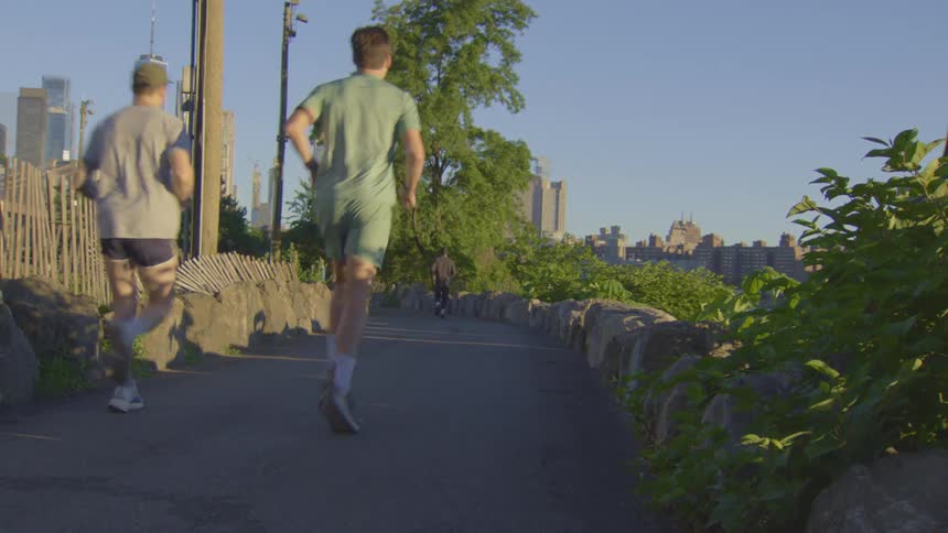A steady shot from behind shows men jogging on a path in a park with the New York City skyline in the background during golden hour
