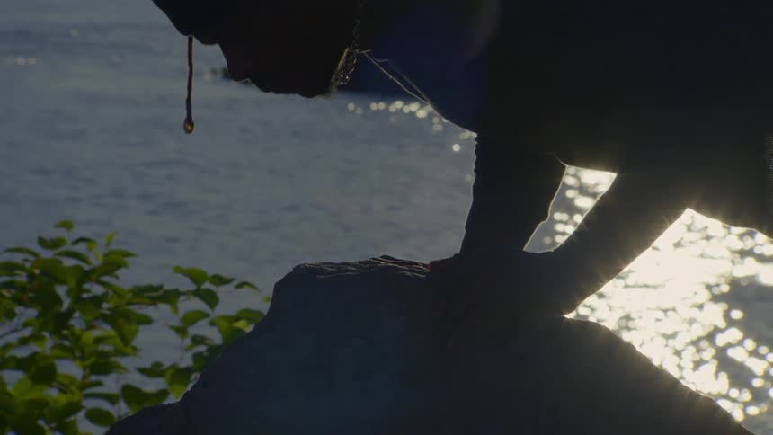 A silhouetted man holds a yoga plank pose on a rock by the water as the sun reflects on the surface