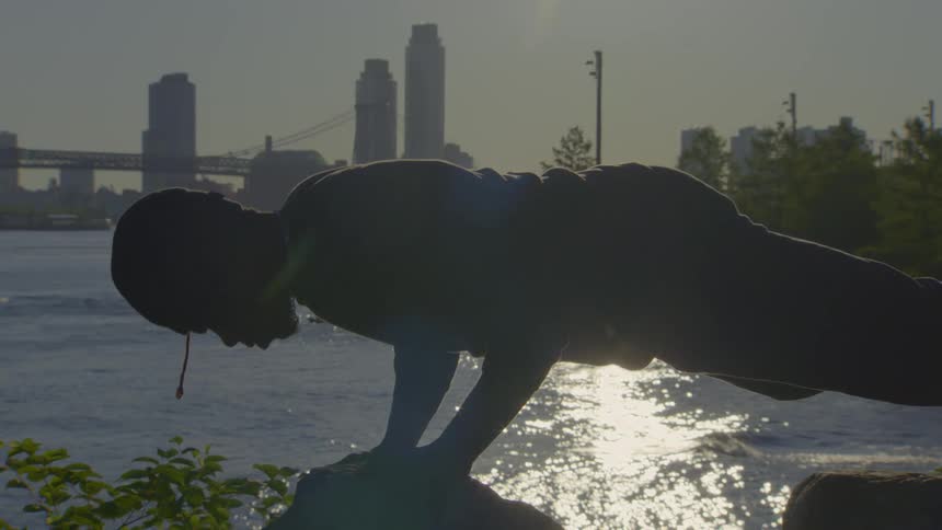 A silhouetted man holds a challenging yoga arm balance pose on a rock by the river at sunrise, with a city skyline in the background